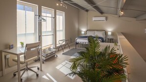 A modern bedroom with a bed, bedside tables, a desk, and a chair. There are large windows allowing natural light, and a ceiling fan is visible.