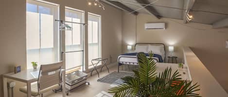 A modern bedroom with a bed, bedside tables, a desk, and a chair. There are large windows allowing natural light, and a ceiling fan is visible.