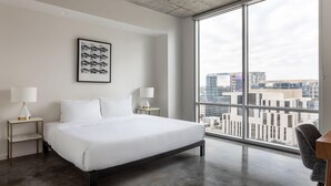 Een moderne hotelkamer met een groot bed, twee nachtkastjes met lampen, een ingelijst schilderij aan de muur en een groot raam met uitzicht op de stad.