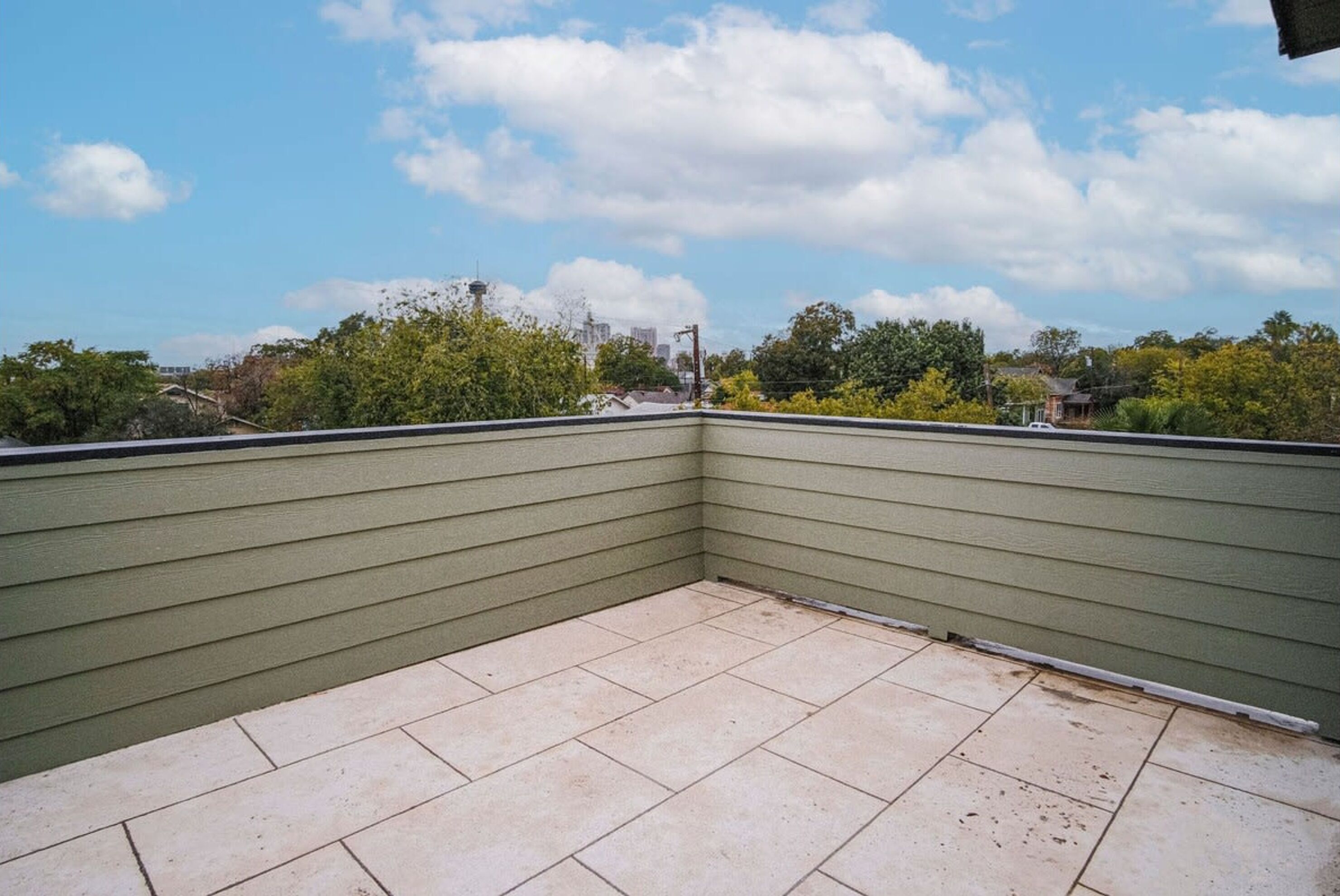 A rooftop terrace with a green exterior wall, tiled floor, and a clear view of the sky and surrounding buildings.