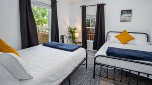 Un dormitorio con dos camas, un ventilador de techo, una ventana con cortinas, una mesita con una lámpara y un cuadro en la pared.