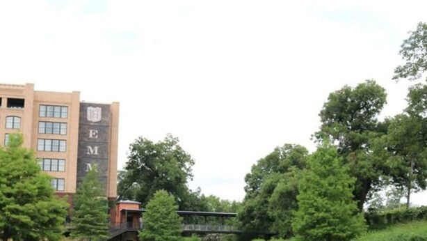 A building with a large 'EM' logo, a calm water body, and lush greenery.