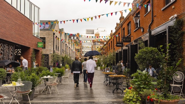 Une rue étroite bordée d’immeubles en briques, avec des terrasses aménagées de tables et de chaises, et des plantes en pot.