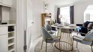 A modern living room with a round dining table, grey upholstered chairs, a blue sofa, and a view of the city through large windows. - The Knightsbridge Suite Next to Sloane Square (London)