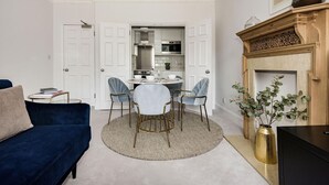 A living room with a blue sofa, a round coffee table, and a dining area with a round table and four chairs. There is a kitchen visible through an open door, and a fireplace with a vase of plants on the right. - The Knightsbridge Suite Next to Sloane Square (London)
