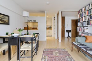 Ein modernes Wohnzimmer mit Essbereich, einer Bücherwand und einer Küche im Hintergrund.