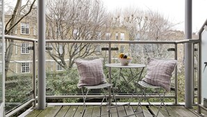 A balcony with two chairs and a table, a bowl of fruit, and a view of buildings and trees. - The Little Library (London)