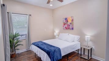 Un dormitorio con cama, lámparas de noche, ventilador de techo, ventana con persianas, planta en maceta y un cuadro abstracto colorido en la pared.