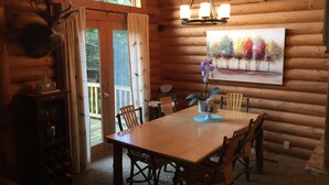 A wooden cabin interior with a dining table, chairs, a chandelier, a painting, and a deer head mounted on the wall. - Quiet cabin in the woods (Traverse City)