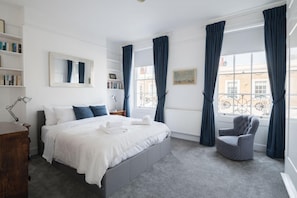 A modern bedroom with a large bed, a chair, and a view of the city through large windows. - At Home in Chelsea (London)