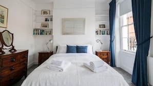 A neatly arranged bedroom with a large bed, two bedside tables with lamps, a dresser, and a bookshelf.