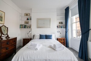 A neatly arranged bedroom with a large bed, two bedside tables with lamps, a dresser, and a bookshelf.