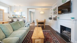 A modern living room with a sofa, a wooden coffee table, a dining area with a table and chairs, a kitchen with cabinets and a sink, and a fireplace with a clock on the wall. - Fabulous Fulham (London)