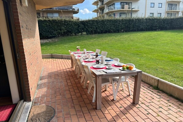 Une terrasse avec une table dressée pour le repas, entourée d’un sol en briques et d’une pelouse verdoyante.