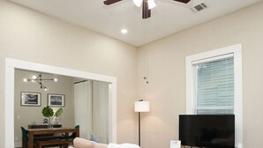 A living room with a beige sofa, a TV on a stand, a ceiling fan, and a dining area in the background. - Amazing Fully Fenced Home Only 1 7 mi to Downtown (San Antonio)