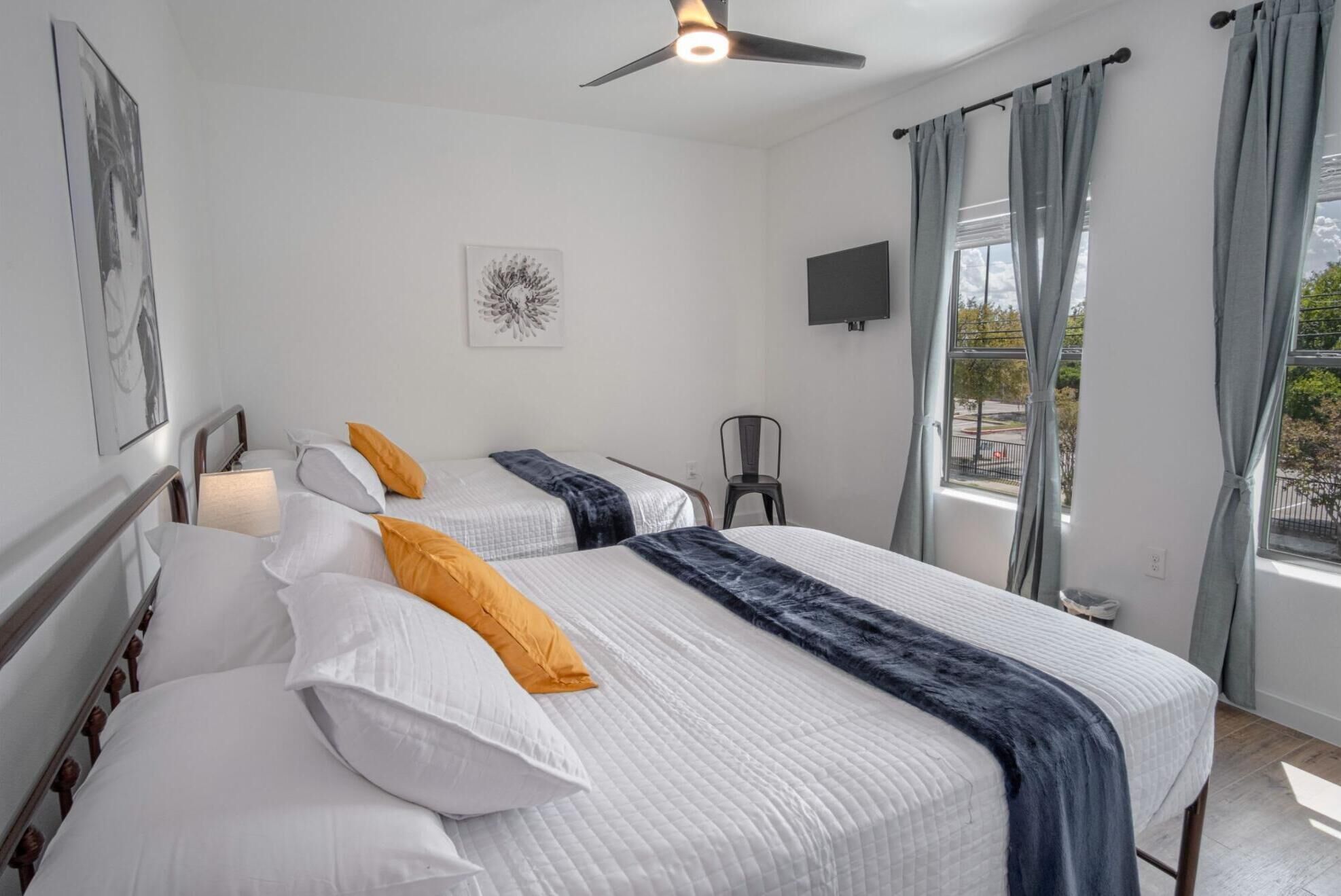 Ein Hotelzimmer mit zwei Betten, einem Deckenventilator, einem an der Wand befestigten Fernseher und einem Fenster mit Vorhängen.