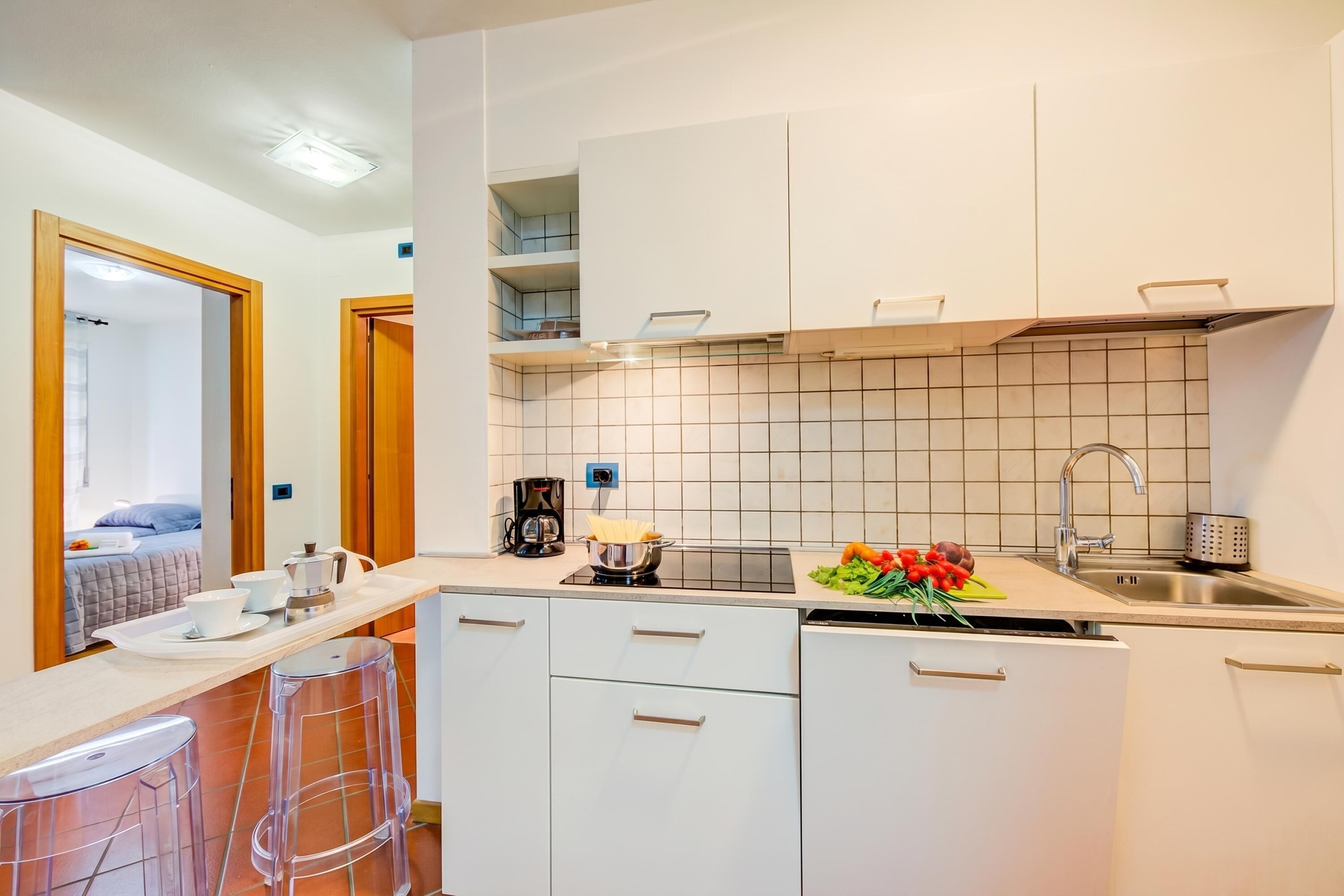 A compact kitchen with white cabinets, a sink, and a countertop with a kettle and a bowl of fruit.