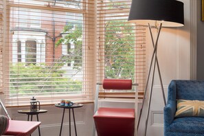 A room with a large window with blinds, a floor lamp, a red stool, a small table with a coffee pot and mugs, and a blue patterned chair. - A West London Retreat (London)