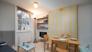 Ett kompakt bostadsutrymme med en matplats, en bokhylla, en öppen spis och ett fönster med utsikt över byggnader.