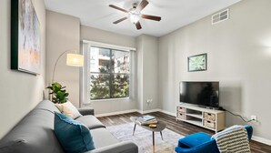 Un salon moderne avec un canapé gris, un fauteuil bleu, un téléviseur à écran plat et un ventilateur de plafond.