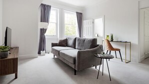 Een moderne woonkamer met een grijze bank, een klein rond tafeltje, een bureau met stoel en een televisie op een houten dressoir.