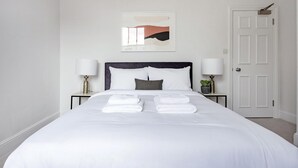 Een net opgemaakt bed met witte lakens en handdoeken, twee nachtlampjes, een lijst met abstracte kunst en een gesloten deur.