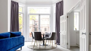 Una habitación con un sofá azul, una mesa redonda con sillas y vistas a edificios a través de un amplio ventanal con cortinas.