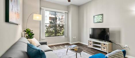 A modern living room with a grey sofa, a blue armchair, a ceiling fan, a flat-screen TV, and a large window.