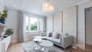 A modern living room with a grey sofa, two white coffee tables, a patterned rug, and a large window with curtains. - Maida Vale Elegance (London)