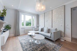 A modern living room with a grey sofa, two white coffee tables, a patterned rug, and a large window with curtains. - Maida Vale Elegance (London)