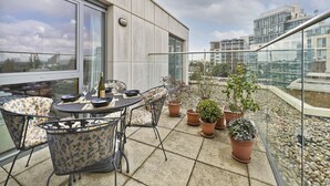 A balcony with a table set for dining, surrounded by potted plants and wicker chairs. - Putney Rooftops (London)