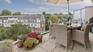 A balcony with wicker furniture, a table, and a large umbrella. There are potted plants and a view of buildings in the background. - Putney Rooftops (London)