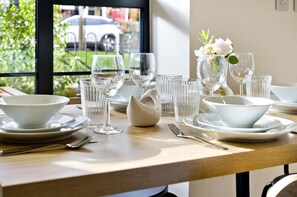 Una mesa de comedor dispuesta con platos, tazones y vasos blancos, y un florero con flores a la derecha.
