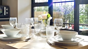 Ein Esstisch ist mit weißen Tellern, Schüsseln, Gläsern und Besteck gedeckt, in der Mitte steht eine Blumenvase.