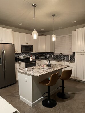 A modern kitchen with white cabinets, a granite countertop, stainless steel appliances, pendant lighting, and a central island with bar stools. - Immaculate guesthouse in the NW Tucson desert...yet close to everything! (Tucson)