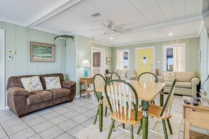Uma sala de estar com um sofá marrom, uma mesa de jantar de madeira com cadeiras, um ventilador de teto e vista para o exterior.
