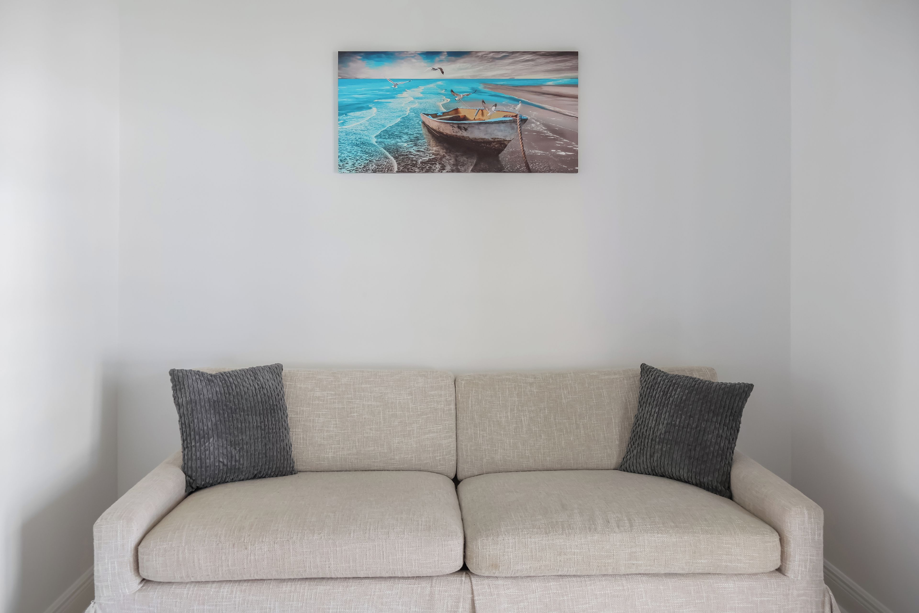 A beige sofa with two grey pillows against a wall, above which is a painting of a boat on a beach.