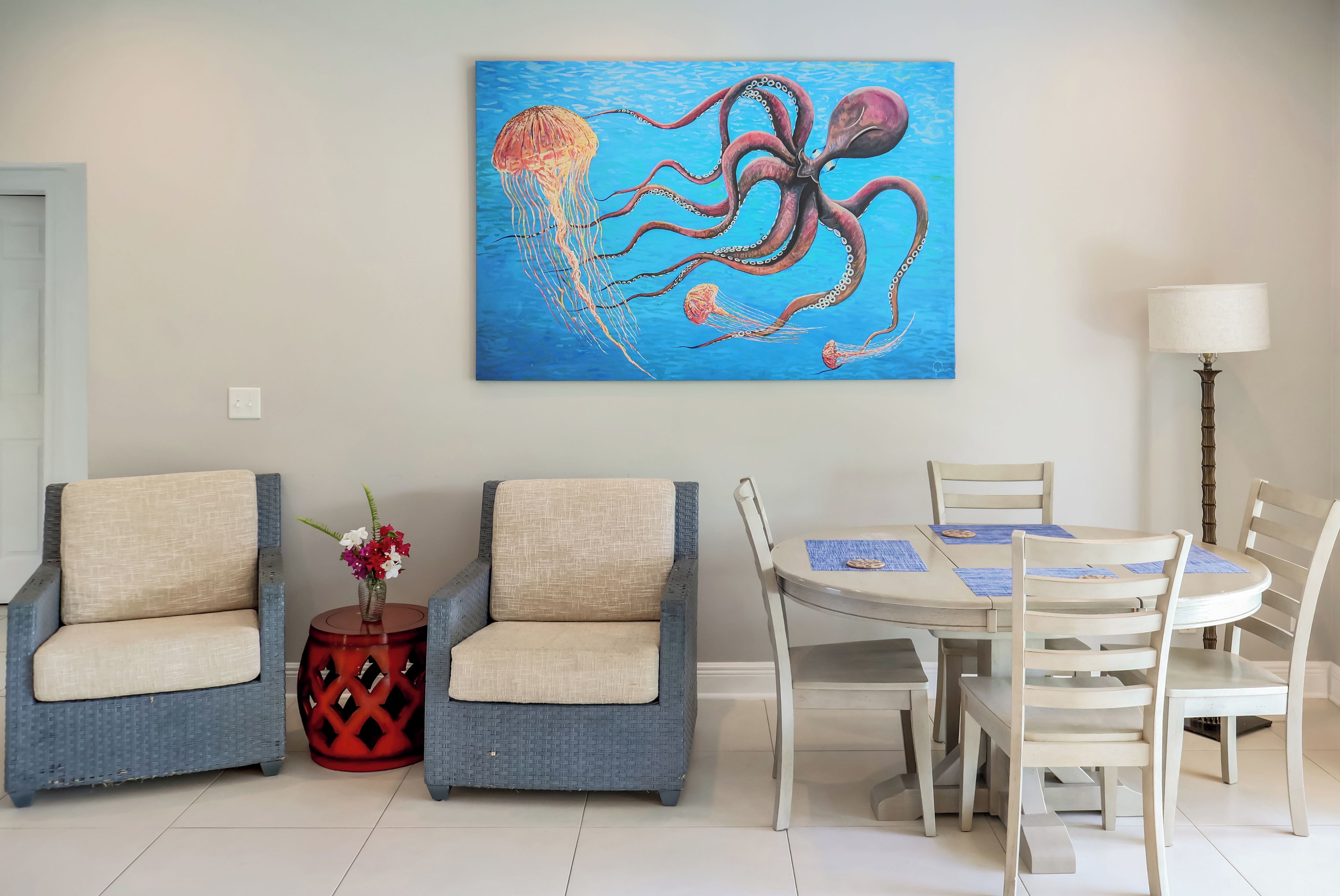 A room with a dining table, two chairs, a red stool, and a wall-mounted painting of an octopus.