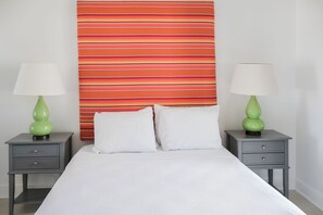 A hotel room with a bed, two nightstands, two green lamps, and a striped headboard. - Colibri Court (Jolly Harbour)