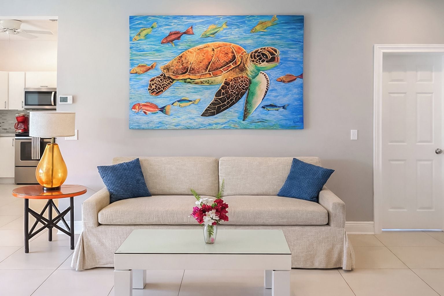 A living room with a beige sofa, a coffee table with a vase of flowers, and a painting of a sea turtle on the wall.