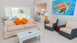 Een woonkamer met een beige bank, twee oranje kussens, een glazen salontafel en een schilderij van een zeeschildpad.