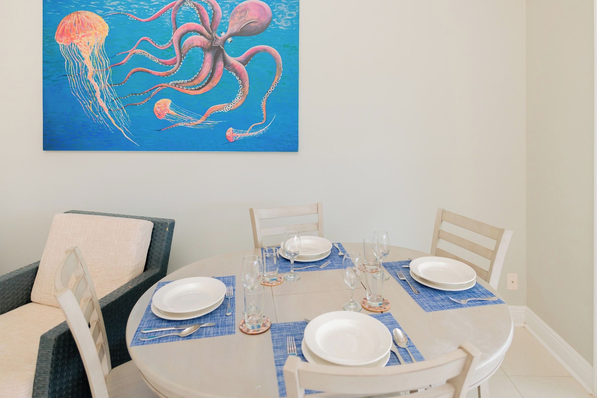 Uma área de refeições com uma mesa redonda posta para quatro pessoas, com pratos brancos, talheres e copos de vinho. Um quadro de polvo e água-viva está pendurado acima da mesa.