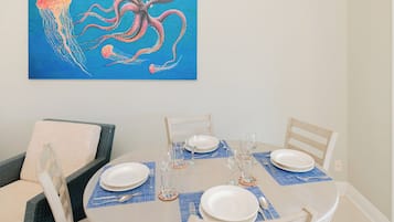 Uma área de refeições com uma mesa redonda posta para quatro pessoas, com pratos brancos, talheres e copos de vinho. Um quadro de polvo e água-viva está pendurado acima da mesa.