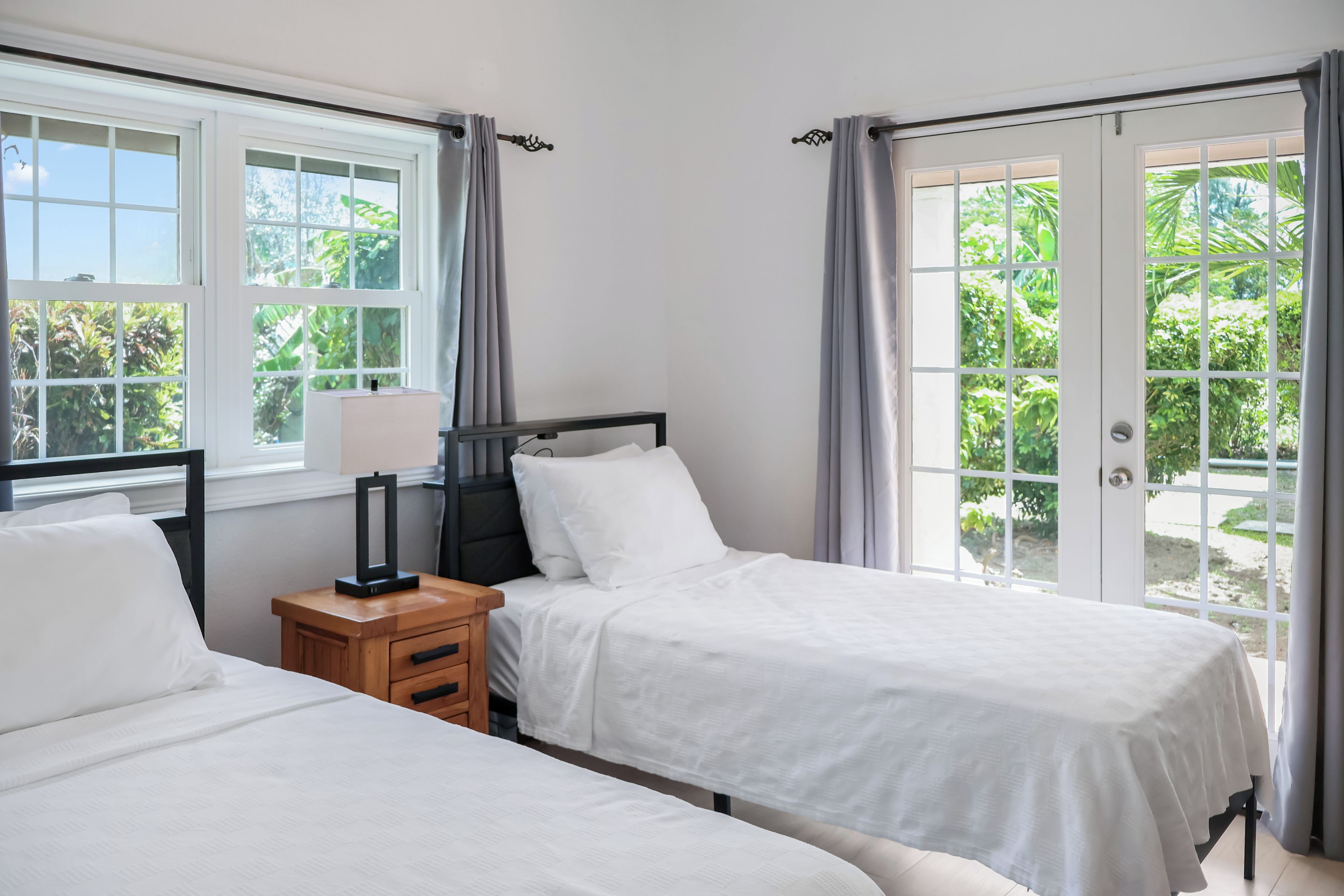 A bedroom with two beds, a bedside table with a lamp, and a view of greenery through the windows.