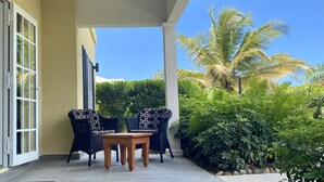 Een terras buiten met twee rieten stoelen en een houten tafel, omgeven door weelderige begroeiing en een palmboom.