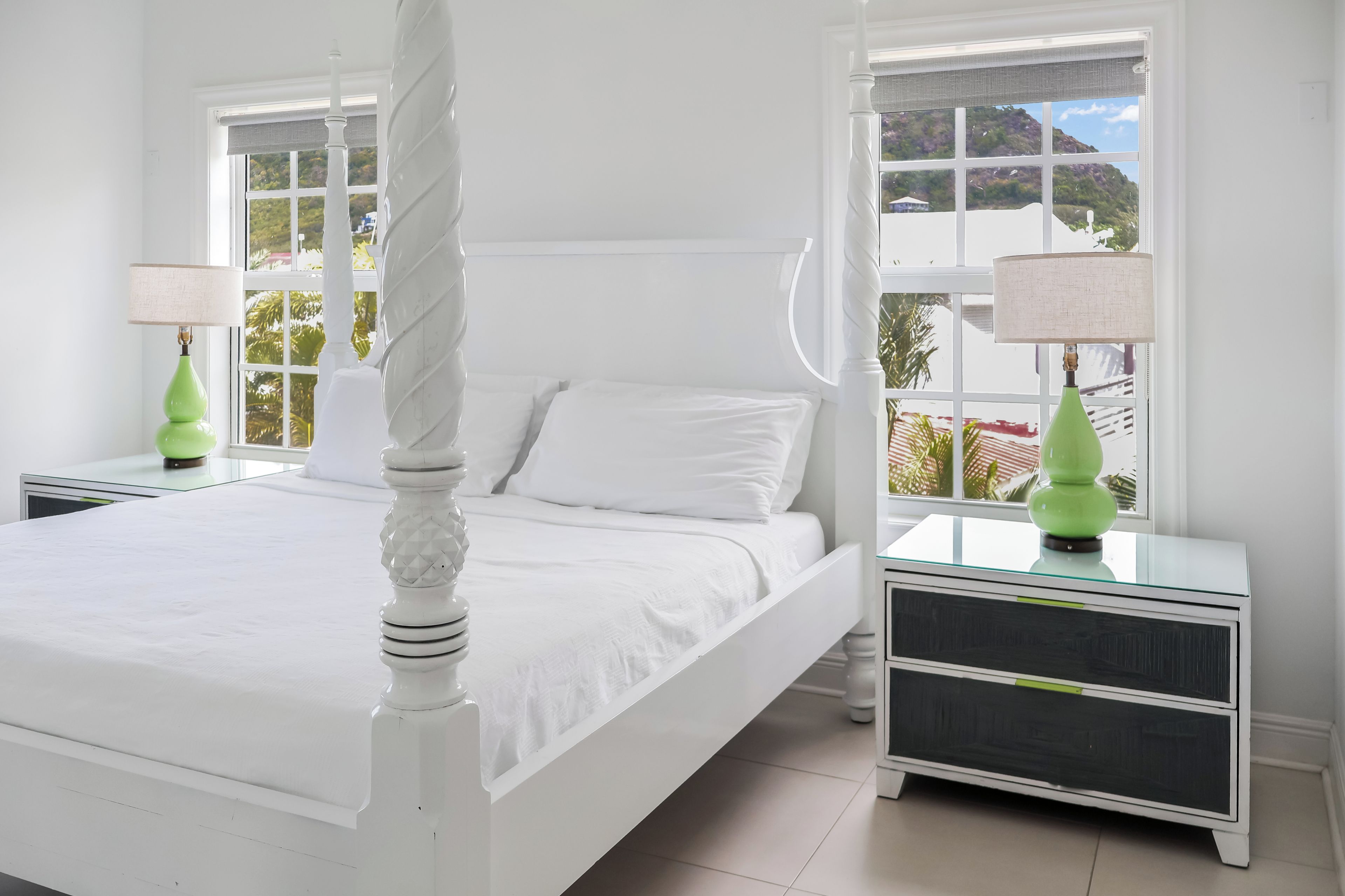 A white four-poster bed with a textured headboard, two nightstands with green lamps, and a view of a mountainous landscape through large windows.