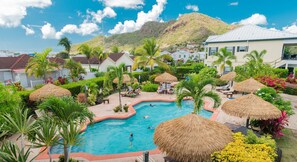 A pool area with thatched umbrellas, palm trees, and a mountain backdrop. - Colibri Court (Jolly Harbour)