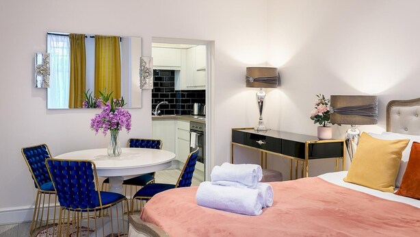 A hotel room with a bed, a round dining table with blue chairs, a small kitchen area, and a mirror. - Designer 1-bedroom in Kensington (London)
