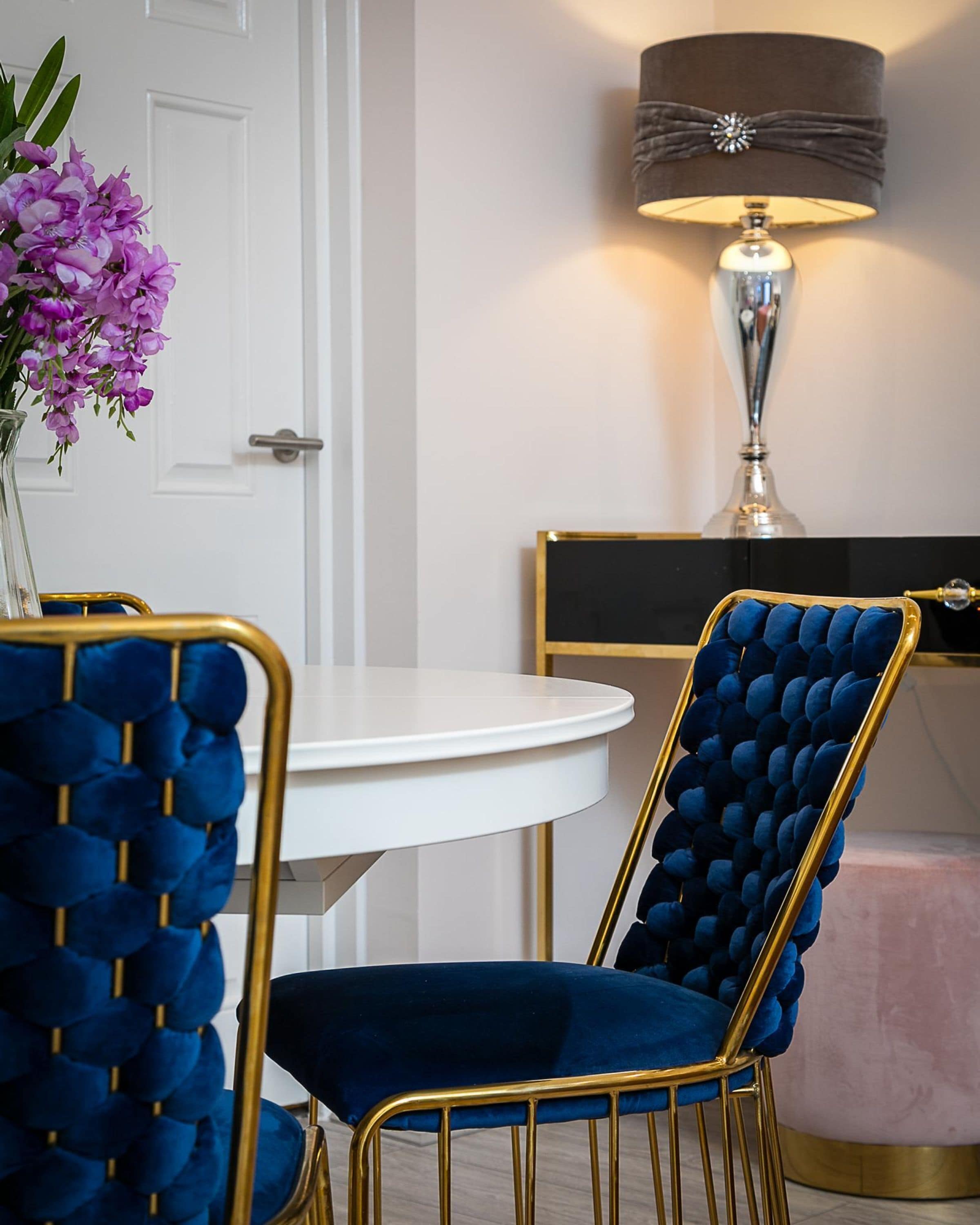 A dining area with a round white table, two blue velvet upholstered chairs with gold frames, a vase with purple flowers, and a wall-mounted lamp.