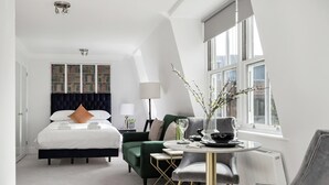 Una habitación de hotel moderna con una cama grande, una zona de comedor con una mesa redonda y vistas de la ciudad a través de amplios ventanales.